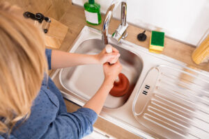 A woman using a plunger in a clogged kitchen sink.